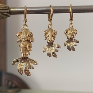 Könnte beinhalten: Goldfarbene, fischförmige Ohrringe hängen an einer Metallstange. Die Ohrringe sind unterschiedlich groß, der größte mit detaillierten Flossen und einem fließenden Schwanz. Einzigartiges Accessoire.