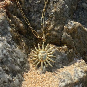 Op de afbeelding: Een goudkleurige ketting met een zonnehanger. De zonnehanger heeft een gezicht in het midden en stralen die naar buiten uitstralen. De ketting wordt getoond op een rotsachtig oppervlak met zand op de achtergrond.