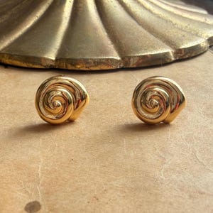 May include: A pair of gold-toned stud earrings featuring a polished, spiralling shell design. Each earring has a smooth, rounded surface that curves inward towards the centre, creating a distinct nautilus-like shape with a reflective metallic finish.
