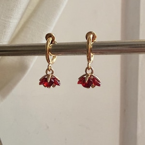 May include: A pair of gold hoop earrings featuring small, dangling red flower-shaped charms. Each charm consists of multiple faceted red petals arranged in a circular pattern, suspended from a thin gold hoop that secures with a small clasp.