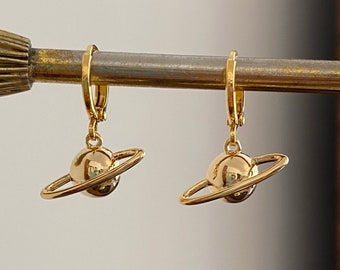 May include: A pair of gold-colored hoop earrings, each featuring a small planet charm with a ring around it. The earrings are hanging on a gold-colored rod. The background is a neutral color.
