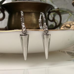 May include: A pair of silver-toned hoop earrings featuring dangling, elongated, pointed cone-shaped charms. The earrings have a smooth, metallic finish and are designed with a hinged closure mechanism at the top of each circular hoop component.