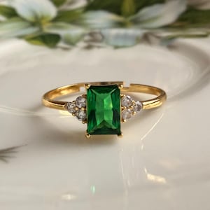 Pode incluir: Um anel de tom dourado com uma pedra central retangular verde-esmeralda, ladeada por três pequenas pedras transparentes de cada lado. A banda é fina e inclui uma abertura ajustável na parte posterior, com acabamento metálico polido.
