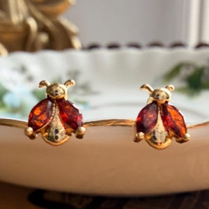 May include: A pair of gold-toned stud earrings shaped like ladybirds, featuring deep red marquise-cut stones for the wings and small gold heads with antennae. The earrings have a polished metallic finish and are displayed in a symmetrical arrangement.