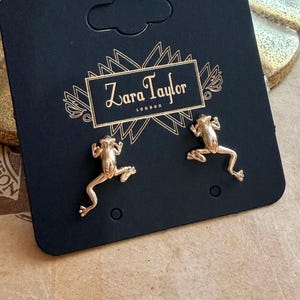 May include: Gold frog earrings displayed on a black card with the text "Zara Taylor London". The earrings are shaped like small frogs. The card is set against a patterned background. The earrings are a delicate piece of jewellery.