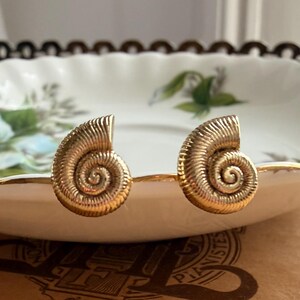 Può includere: Un paio di orecchini color oro a forma di conchiglie di nautilo a spirale. Ogni orecchino presenta una superficie testurizzata e scanalata che segue la curva della conchiglia, creando un distinto motivo avvolto. Finitura metallica lucida.