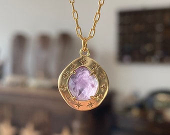 May include: A gold-coloured necklace with a pendant featuring a light purple gemstone. The pendant is surrounded by a gold frame with star-shaped engravings. The chain is also gold-coloured.