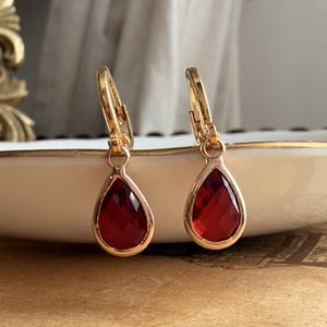 Peut inclure: Une paire de boucles d'oreilles créoles dorées avec des pierres rouges en forme de goutte à facettes suspendues. Les boucles d'oreilles présentent un mécanisme de fermeture à charnière en haut de l'anneau et un sertissage en métal doré poli.