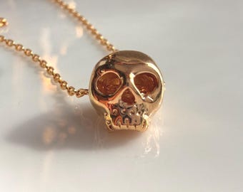 Könnte beinhalten: Eine goldfarbene Metallkette mit einem kleinen, dreidimensionalen Totenkopf-Anhänger. Der Totenkopf hat hohle Augenhöhlen und einen detaillierten Kiefer. Der Anhänger ist an einer zarten, passenden goldfarbenen Kette befestigt, die sich durch das Bild zieht.