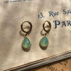 Könnte beinhalten: Ein Paar goldene Creolen mit tropfenförmigen grünen Edelsteinanhängern. Die Ohrringe sind auf einem alten Buch mit französischem Text abgebildet.