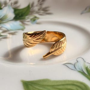 Puede incluir: Un anillo abierto de tono dorado que presenta dos extremos superpuestos en forma de hoja. La banda tiene una superficie pulida y reflectante con detalles de venas grabados en cada hoja. El anillo está diseñado con un espacio entre las hojas.