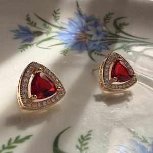 May include: A pair of gold-tone stud earrings featuring a triangular-cut red gemstone at the centre, surrounded by a halo of small, clear pavé-set stones. The earrings have a metallic gold-coloured frame and a post-back closure mechanism.