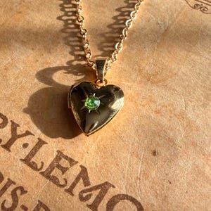 May include: A gold-toned heart-shaped locket pendant suspended from a matching link chain. The locket features a small, round, light green gemstone set in the centre, surrounded by an engraved starburst pattern on the polished metal surface.