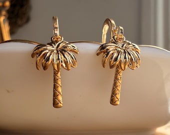 May include: A pair of gold-toned hoop earrings featuring dangling palm tree charms. Each charm is detailed with a textured trunk and radiating fronds, suspended from a small circular hoop mechanism designed for attachment to the earlobe.