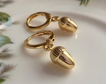 Puede incluir: Un par de pendientes de aro dorados con colgantes en forma de bellota. Cada colgante tiene una base lisa y pulida y una tapa texturizada con un patrón de líneas cruzadas. Los pendientes cuentan con un cierre de bisagra para mayor seguridad.