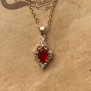 Puede incluir: Un collar de cadena dorada con un colgante de piedra roja. El colgante está rodeado de piedras transparentes más pequeñas y tiene un diseño detallado y ornamentado.