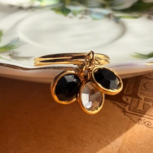 Pode incluir: Um anel de tom dourado com três pingentes ovais pendurados. Dois dos pingentes contêm pedras pretas opacas, enquanto o pingente central apresenta uma pedra transparente facetada. O aro do anel é liso e metálico, com todos os elementos em ouro.