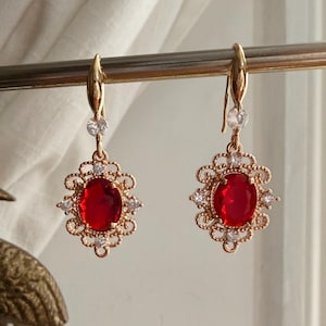 Puede incluir: Un par de pendientes dorados con una piedra central roja de forma ovalada, rodeada de pequeñas piedras blancas y un diseño de filigrana dorada ornamentada. Los pendientes cuelgan de un gancho dorado.