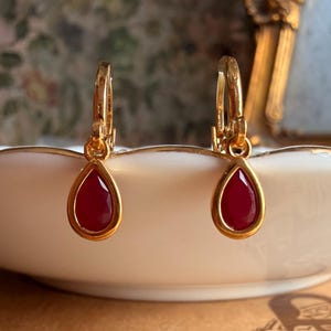 May include: A pair of gold-toned hoop earrings featuring dangling teardrop-shaped charms with smooth, deep red centers. The earrings have a polished metallic finish and a secure hinged closure mechanism, presenting a classic and symmetrical design.