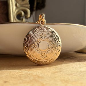 May include: A round gold-tone metal locket pendant featuring an intricate embossed pattern on its surface. The locket has a central circular design surrounded by a textured grid and scalloped edges, with a small bail at the top for a chain.