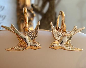 Könnte beinhalten: Ein Paar goldfarbene Creolen, die jeweils mit einem hängenden Schwalbenanhänger versehen sind. Die Vögel sind im Flug mit ausgebreiteten Flügeln dargestellt und weisen detaillierte Linien auf den Flügeln und Schwanzfedern auf. Die Creolen sind rund.