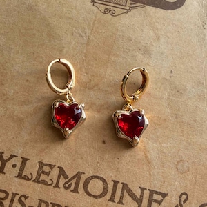May include: Two gold-coloured hoop earrings, each featuring a heart-shaped, red gemstone pendant. The earrings are set against a vintage paper background with printed text.