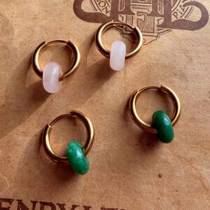 May include: Four gold-colored hoop earrings, each with a small, colorful stone. Two earrings have light pink stones, and two have green stones. The earrings are displayed on a vintage sheet of music with text.