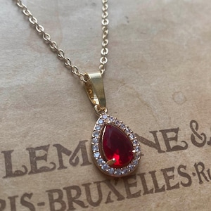 Può includere: Collana dorata con un pendente a forma di goccia. Il pendente presenta una grande gemma rossa circondata da piccole pietre trasparenti. La catena è delicata e il pendente è appeso a una semplice cauzione.
