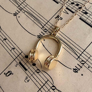 Puede incluir: Un collar dorado con un colgante en forma de auriculares. El colgante cuelga de una fina cadena. El fondo es una página de partituras vintage con notas y símbolos musicales.