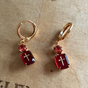Può includere: Un paio di orecchini a cerchio color oro con un ciondolo composto da un piccolo cristallo rosa rotondo e un cristallo rosso intenso rettangolare più grande. Ogni cristallo rosso presenta un piccolo emblema a croce dorato al centro.