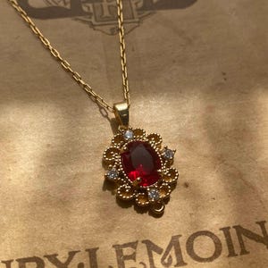 Peut inclure: Un collier doré avec un pendentif orné d'une grosse pierre rouge ovale entourée de petites pierres claires. Le pendentif est suspendu à une fine chaîne dorée. Le fond est un papier vintage.