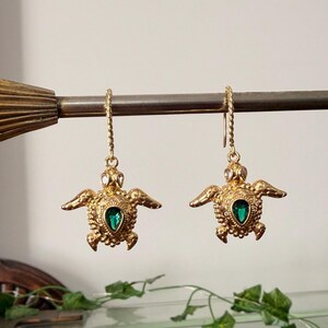 May include: A pair of gold-colored turtle earrings with green gemstone centers. The earrings are hanging from a metal rod. The turtles have outstretched wings and detailed textures. The background includes a decorative brass fixture and a vintage display case.