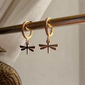 May include: A pair of gold-toned hoop earrings, each featuring a small, dangling dragonfly charm. The earrings are suspended from a thin, horizontal metal bar, showcasing a polished metallic finish and a simple, symmetrical insect design.