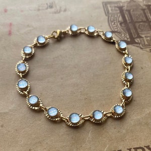 以下が含まれることがあります： 繊細なチェーンと丸い白くオパールのような石がついた金のブレスレット。ブレスレットは、ヴィンテージ風の紙の背景に置かれており、「HENRY-LE 17-Rue Pigalle (IX) PARIS」というテキストが印刷されています。