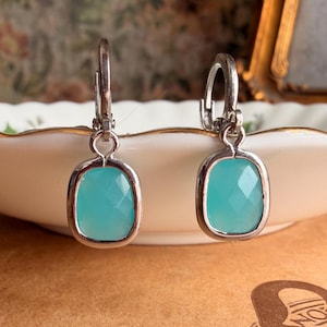 Puede incluir: Un par de pendientes colgantes con piedras de color azul claro talladas en forma cuadrada y engastadas en marcos de metal plateado. Los pendientes cuentan con cierres de palanca y un acabado pulido que resalta el tono azul vibrante.