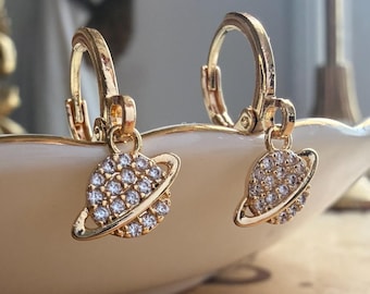 May include: A pair of gold-toned hoop earrings featuring a dangling planet-shaped charm. Each charm is encrusted with small, clear, sparkling stones and includes a ring encircling the center, resembling the planet Saturn in a metallic finish.