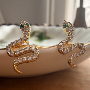 Könnte beinhalten: Ein Paar goldfarbene Ohrstecker in Schlangenform mit einem gewundenen Körper, der mit klaren weißen Kristallen besetzt ist, sowie zwei kleinen grünen Steinen als Augen. Die Ohrringe verfügen über einen Stiftverschluss und ein glänzendes Finish.