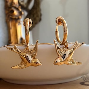May include: A pair of gold-toned hoop earrings, each featuring a dangling charm shaped like a flying swallow bird. The earrings have a polished metallic finish with detailed engravings on the wings of the birds, suspended from circular hoops.