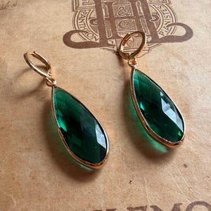 Puede incluir: Un par de pendientes colgantes con piedras de color verde esmeralda en forma de lágrima facetada, engastadas en marcos de metal dorado. Cada pendiente cuelga de un cierre de aro articulado dorado, mostrando un acabado pulido en las gemas.