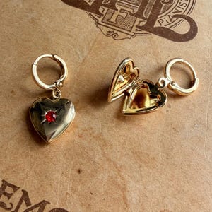 May include: Two gold-toned heart-shaped locket earrings with hoop attachments. One earring is closed and features a small, centered red gemstone. The second earring is open, revealing a hollow interior space within the gold-toned metal heart.
