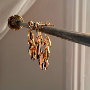 May include: Gold-toned leaf earrings with a post-back closure. The earrings feature multiple dangling leaves in varying shades of gold and rose gold. The earrings are displayed on a metal rod against a soft, neutral background.