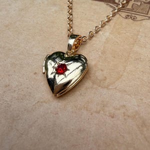 Könnte beinhalten: Goldfarbene herzförmige Medaillon-Halskette mit einem kleinen roten Edelstein in der Mitte. Das Medaillon hängt an einer feinen Goldkette. Der Hintergrund ist ein Papier im Vintage-Stil mit verblasstem Text und Illustrationen.