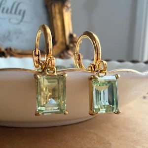May include: A pair of gold hoop earrings featuring rectangular, emerald-cut light green gemstones. Each earring has a polished gold metal hoop with a hinged closure and a dangling, faceted green stone set in a simple four-prong gold frame.
