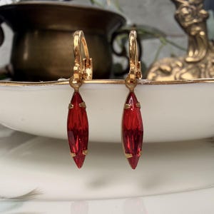 Peut inclure: Une paire de boucles d'oreilles à fermoir levier doré avec des pierres en verre rouge de forme marquise allongée. Chaque cristal rouge vif est maintenu par une monture métallique simple, suspendu à un anneau doré poli pour oreilles percées.