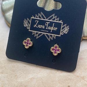 May include: Gold-coloured stud earrings featuring a four-leaf clover design, each set with a cluster of small, pink gemstones. The earrings are presented on a navy blue card with the brand name "Zara Taylor London" in gold lettering.