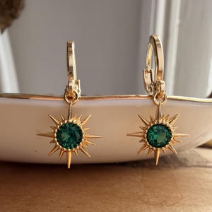 Puede incluir: Un par de pendientes de aro dorados con un colgante en forma de estrella. Cada colgante presenta una piedra preciosa redonda y facetada de color verde esmeralda rodeada de puntas metálicas radiantes que crean una silueta de estrella.