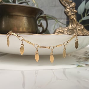 Op de afbeelding: Een goudkleurige kettingarmband met meerdere kleine, platte, bladvormige bedels die aan de schakels hangen. De armband is opgebouwd uit een reeks onderling verbonden metalen schakels en wordt gepresenteerd in een gebogen witte keramische schaal.