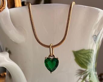 May include: A gold-toned snake chain necklace featuring a small, faceted heart-shaped emerald green pendant. The pendant is suspended from a simple gold-toned bail, showcasing a vibrant green gemstone set against the metallic gold chain.
