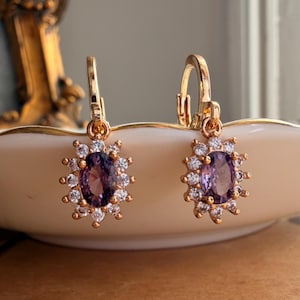 May include: A pair of gold-toned leverback earrings featuring a central oval-cut purple gemstone surrounded by a halo of small, clear, round-cut white stones arranged in a sunburst pattern, creating a bright and reflective appearance.