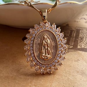 Peut inclure: Un collier pendentif ovale doré présentant une figure centrale en relief de la Vierge de Guadalupe. Le pendentif est entouré d'un halo de pierres de zircone cubique transparentes taille baguette disposées en rayons, suspendu à une chaîne dorée.
